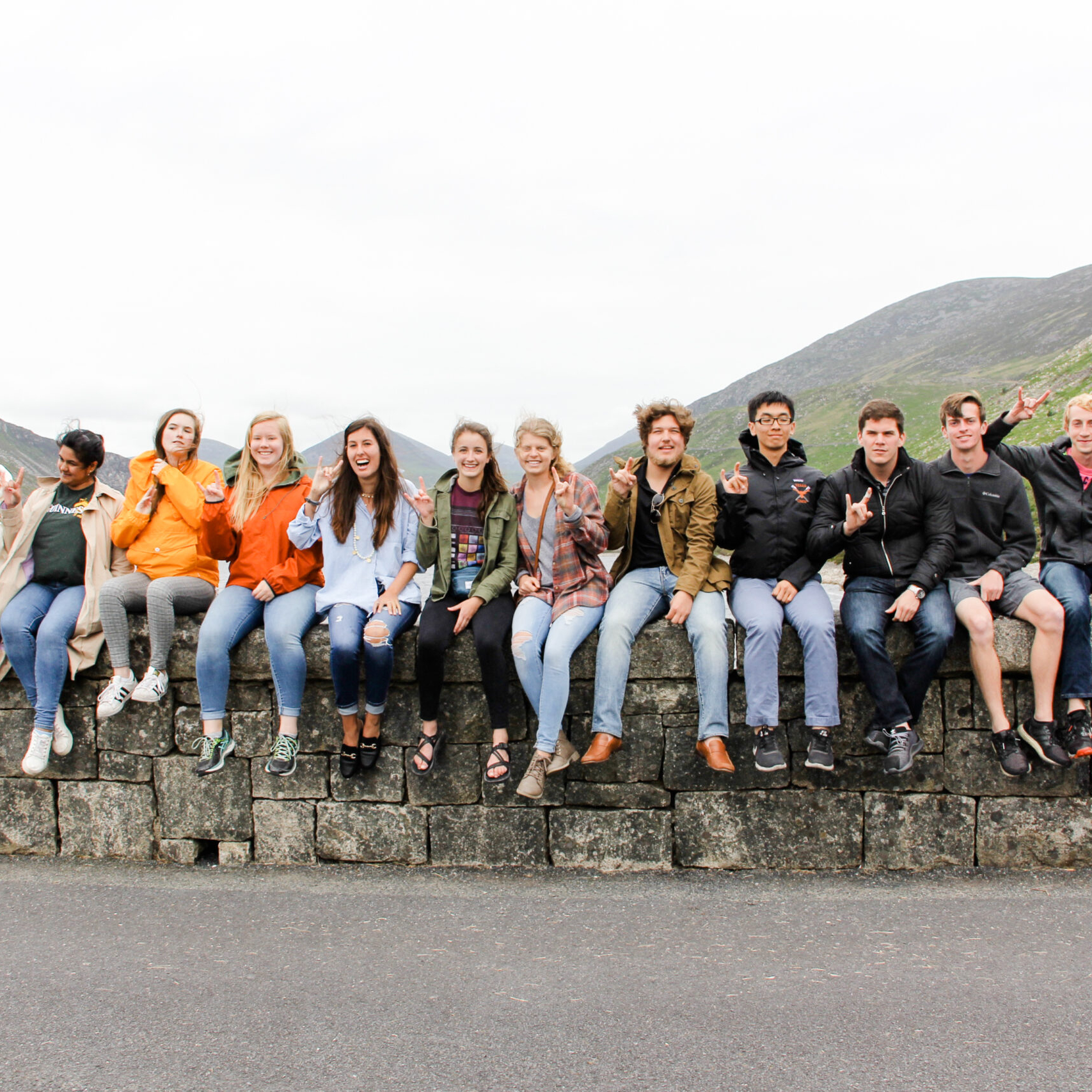 Study abroad students sit on a stone wall, smiling with horns up hand signal