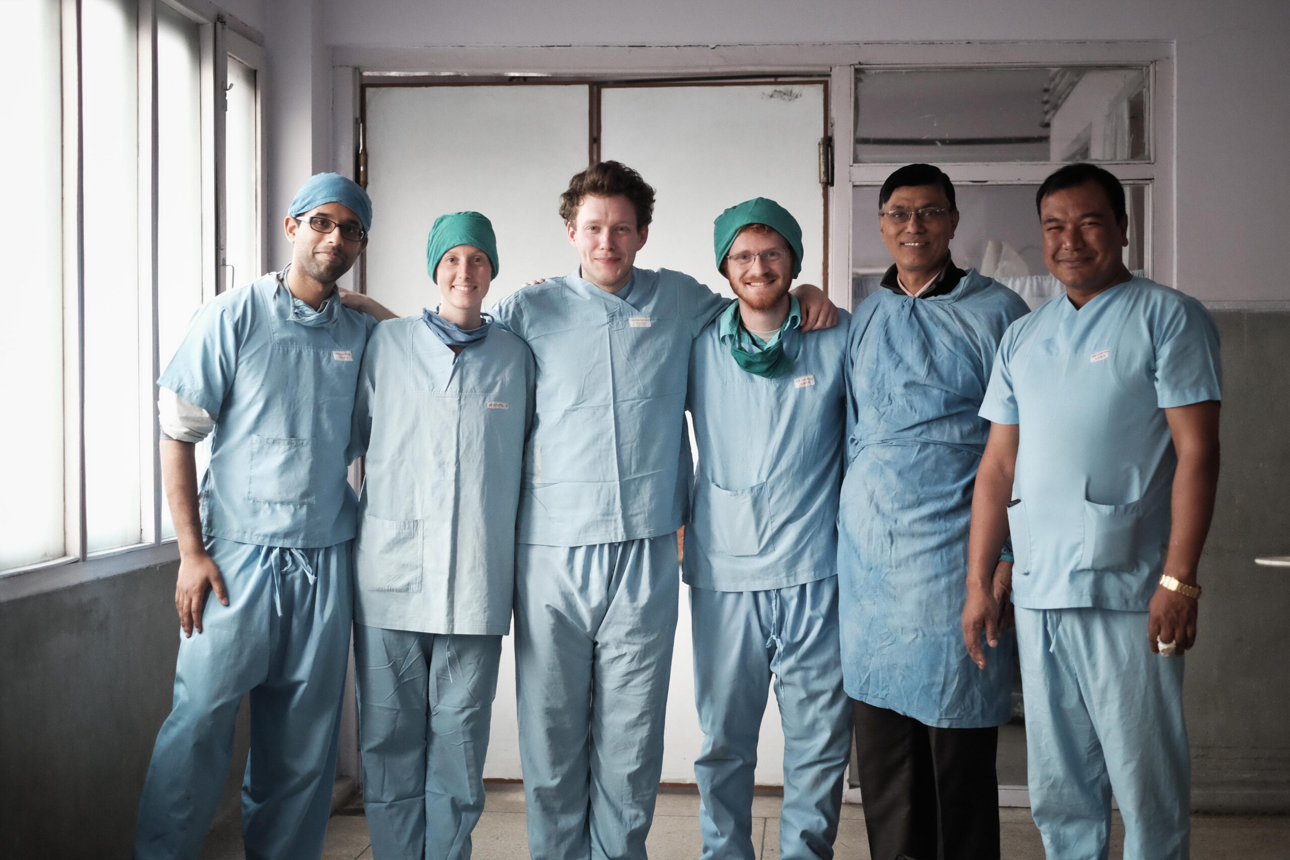 Six people pose in a row, wearing scrubs in a hospital, with arms around each other