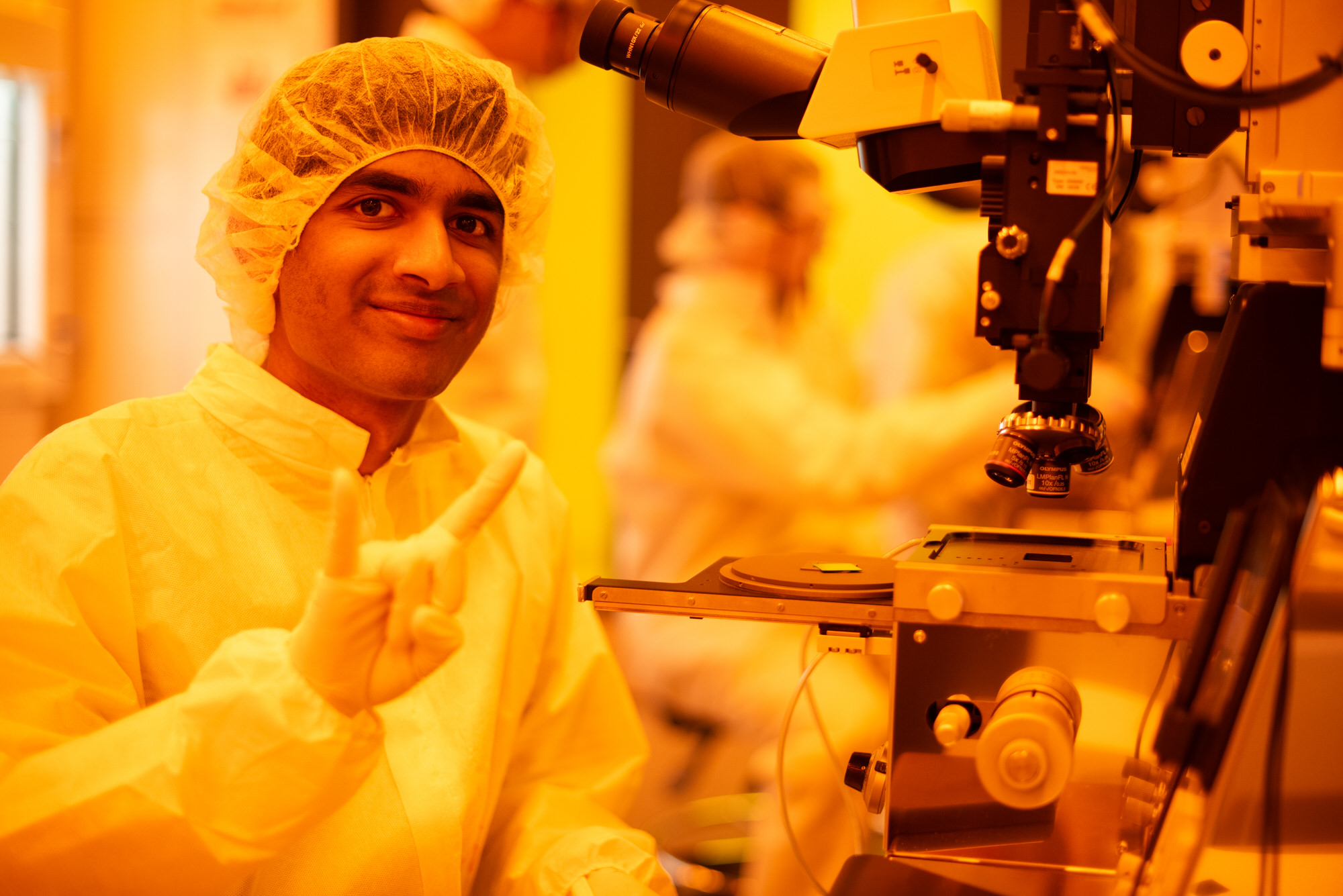 Texas Engineer dressed in lab gear doing hook 'em horn hand sign next to large microscope