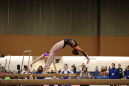 Gymnast performing back handspring on balancing beam in front of crowd