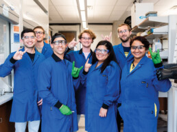 Texas Engineering graduate students in Joan Brennecke lab doing hook 'em hand signs in lab while wearing lab safety gear